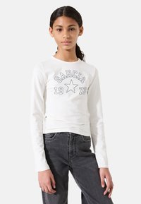 Witte blouse met lange mouwen met de tekst "GARCI A 1972" en een stergrafiek; gecombineerd met strakke donkergrijze jeans. Soepele textuur en casual ontwerp.