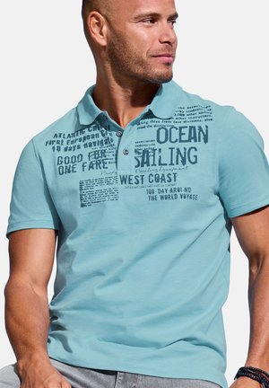 Man wearing a light blue polo shirt with dark blue sailing-themed text and graphics, looking to the right.