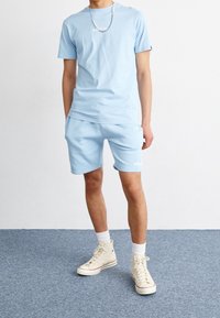 Homme portant un t-shirt ellesse bleu clair et un short assorti, des baskets montantes blanches et un collier de perles, debout sur un tapis bleu.