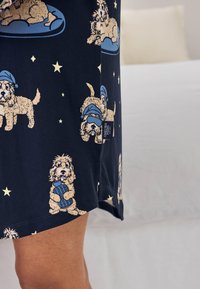 Marineblåt stof med illustrationer af små hunde iført natluer, der holder varmeflasker, og stjerner, vist på tøj nær knæet.