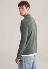 Zeleni pleteni pulover z teksturiranim vzorcem, dolgimi rokavi in prilagojenim robom, kombiniran s svetlo modrimi denim kavbojkami.