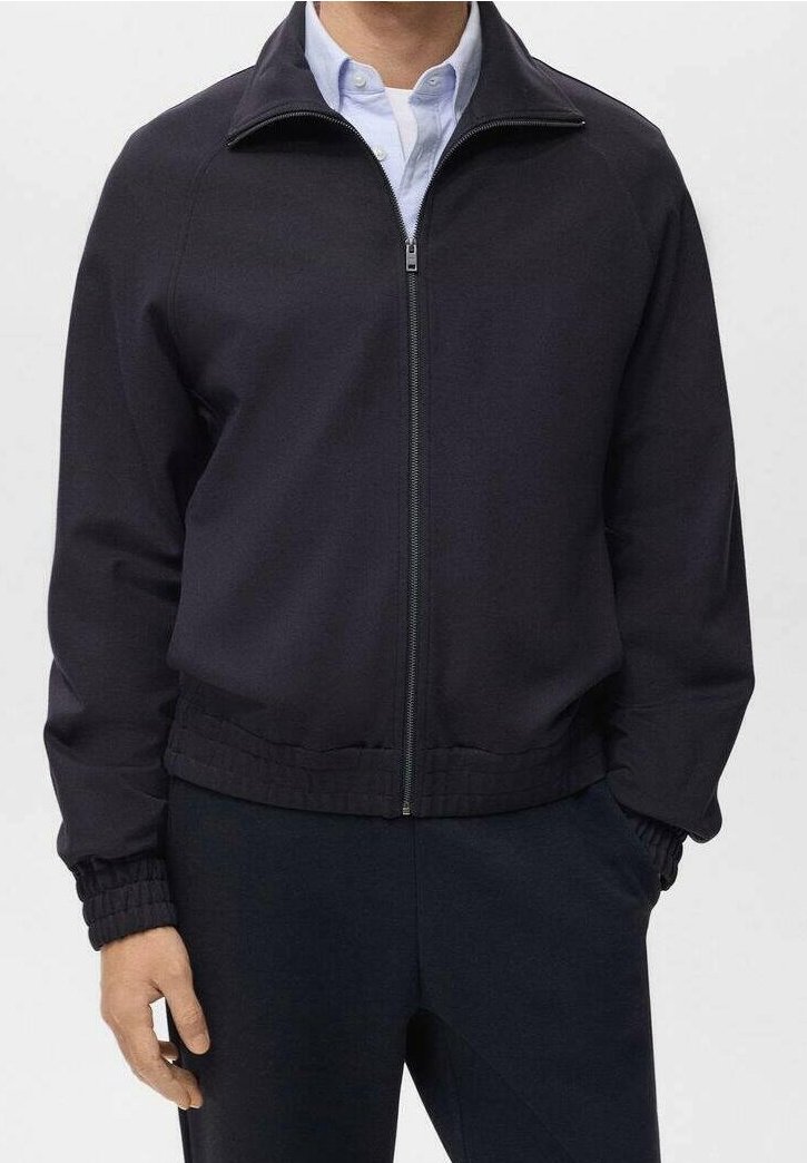 Homme portant une veste noire zippée par-dessus une chemise bleu clair à col, les mains dans les poches, montré du cou aux hanches.