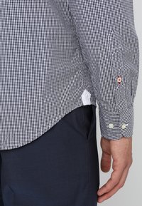 Černobílá kostkovaná košile s bílým lemováním, má kontrastní knoflíky na manžetách a zakřivený spodní lem.