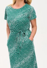 Green dress made of soft fabric with a white hearts pattern, short sleeves, a round neckline, and a tied waist with pockets.