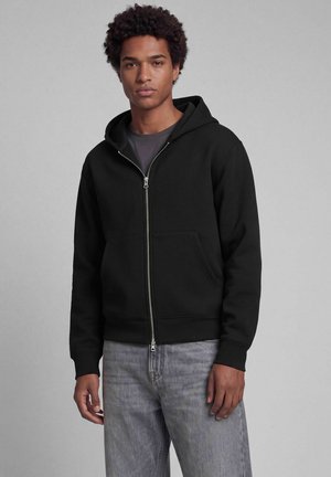 Jonge man met krullend haar die een zwarte hoodie met rits en grijze spijkerbroek draagt, staand voor een effen grijze achtergrond.