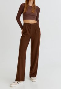 Vrouw in hoge taille, bruine wijde broek, een aansluitend geribbeld langemouwen top en beige sneakers, staande tegen een effen achtergrond.