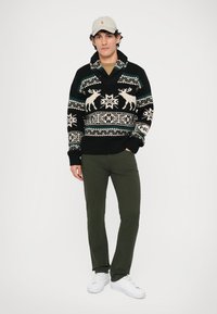 Μαύρο πλεκτό πουλόβερ με λευκά και teal χειμωνιάτικα μοτίβα, με γιακά σαλονιού. Συνδυασμένο με ελιές πράσινα παντελόνια και λευκά αθλητικά παπούτσια.