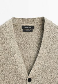 Beige knitted cotton cardigan with black button and visible Massimo Dutti brand label inside the collar.