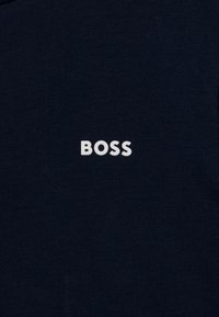 Námořnická modrá tričko vyrobené z bavlny; na přední straně se nachází bílá logo "BOSS" vytištěná tučným písmem bezpatkového typu. Hladký povrch.
