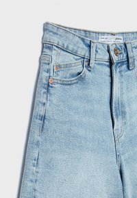 Vaqueros de mezclilla azul claro con cintura alta, diseño de cinco bolsillos y detalles en hardware de cobre. Textura desgastada cerca de la costura lateral.