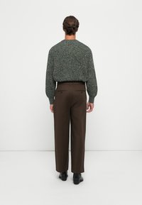 Man seen from behind wearing a green textured sweater, dark brown trousers, and black shoes standing against a plain light background.