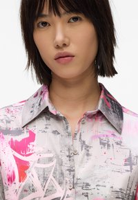 Ovratna srajca z sivo-rožnatim abstraktnim vzorcem, ki ima gumbe in logo na prsih, izdelana iz gladkega, sijočega materiala.