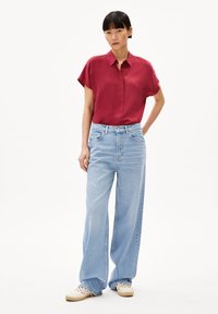 Rode korte mouwen overhemd met knopen, lichtblauwe wijde jeans en geweven witte schoenen. De outfit heeft een ontspannen pasvorm en zachte texturen.