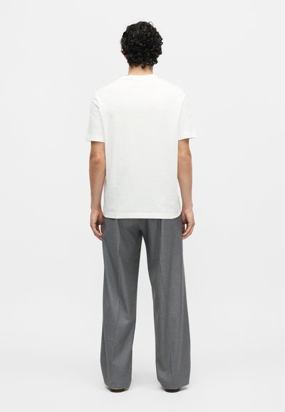 Άνδρας με μαύρα σγουρά μαλλιά στέκεται με την πλάτη, φορώντας ένα απλό λευκό κοντομάνικο t-shirt και άνετο γκρι παντελόνι σε ανοιχτό φόντο.
