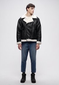 Zwarte leren jas met witte schapenvachtkraag en manchetten, ritssluiting, gecombineerd met lichtblauwe spijkerbroek en zwarte laarzen, op een neutrale achtergrond.