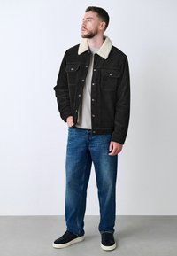Chaqueta de pana negra con cuello de borreguito crema, dos bolsillos frontales, combinada con jeans azules y zapatillas negras, de pie contra un fondo claro.