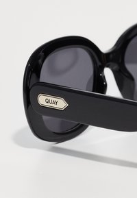 Mustad ümmargused mustad prillid tumedate läätsedega ja kuldne Quay logo silt peaasja küljes valge tausta ees.