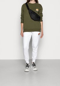 Felpa verde oliva con scollatura rotonda, abbinata a pantaloni jogger bianchi con logo arancione. Borsa a tracolla nera e sneakers chunky.