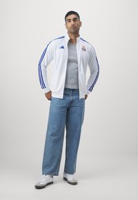 Hvit zip-up jakke med blå striper, med logo, parret med lyseblå vide jeans og hvite sneakers, på en ensfarget bakgrunn.