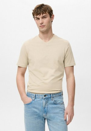 Junger Mann trägt beige V-Ausschnitt T-Shirt und hellblaue Jeans, steht mit einer Hand in der Tasche vor weißem Hintergrund.