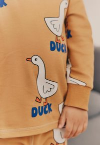 Haut à manches longues de couleur moutarde avec des graphiques de canard blancs ; comporte un texte bleu indiquant "CANARD" ; tissu en coton lisse, poignets côtelés.