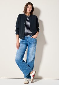 Giubbotto bomber blu scuro sopra una maglietta a righe bianche e blu navy, abbinato a jeans a gamba larga blu chiaro e sneakers bianche con dettagli rossi.