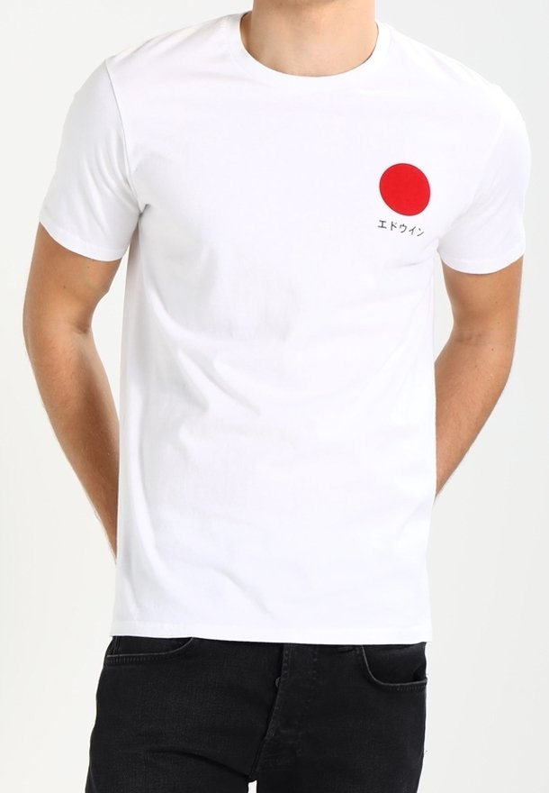 T-shirt blanc à manches courtes avec un cercle rouge et un texte japonais sur le côté gauche de la poitrine, porté par une personne avec un pantalon noir.