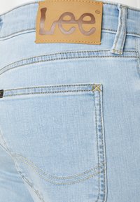 Světlé modré džíny vyrobené z denimu s koženou nášivkou s logem "Lee", žlutým prošitím a elegantním designem, který zdůrazňuje oblast zadní kapsy.