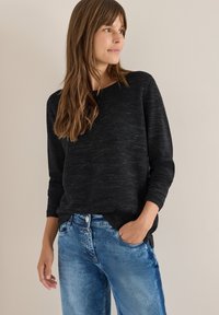 Zwarte gebreide trui met driekwartmouwen, gecombineerd met blauwe denim jeans. De trui heeft een losse pasvorm en een geribbelde onderrand.