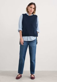 Marineblauwe mouwloze trui over een lichtblauw gestreept overhemd met knopen, gecombineerd met blauwe jeans en donkerrode ballerina's. Eenvoudige, casual lagen.