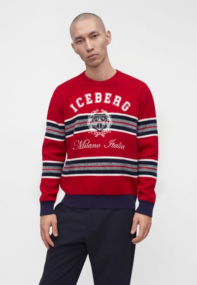 Homme portant un pull rouge avec des rayures blanches et bleu marine, texte "ICEBERG Milano Italia" et emblème, assorti à un pantalon bleu marine, debout devant un fond uni.