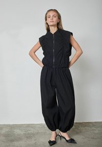 Čierny overal so zipsom vpredu, krátkymi nariasenými rukávmi a voľnými nohavicami dĺžky po členky. Nosí sa s čiernymi sandálmi na remienky.