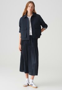 Marineblauwe jas met twee voorzakken, gecombineerd met een geplisseerde donkere rok en witte sneakers. Rechte snit, soepel materiaal en casual ontwerp.