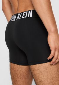 Crne pamučne elastične bokserice s bijelim pojasom s logom "Calvin Klein", koje imaju uski kroj i glatku teksturu.