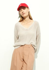 Light beige knitted sweater with a V-neck, paired with light pink trousers. The model wears a red baseball cap with a white logo.