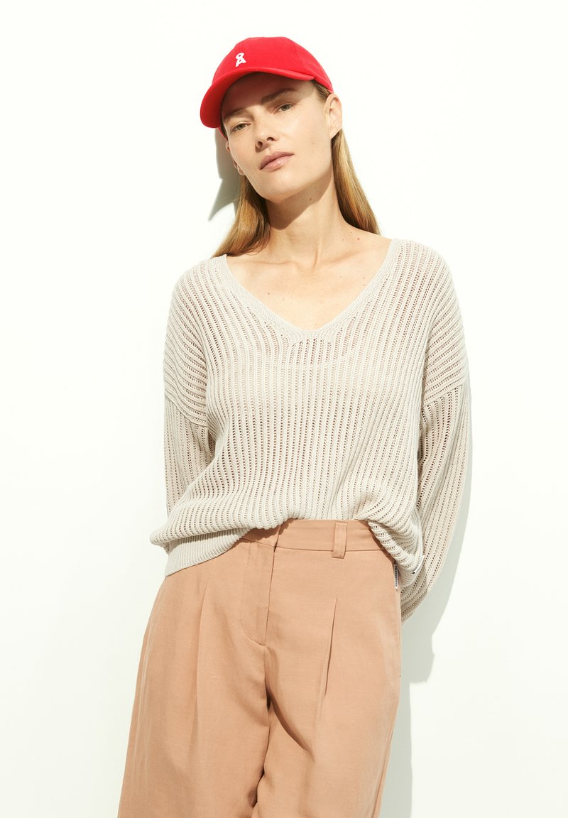 Light beige knitted sweater with a V-neck, paired with light pink trousers. The model wears a red baseball cap with a white logo.
