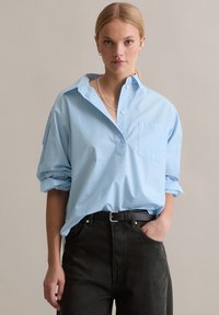 Camicia azzurro chiaro oversize con colletto e taschino sul petto, abbinata a jeans verde scuro e cintura nera, con maniche arrotolate.