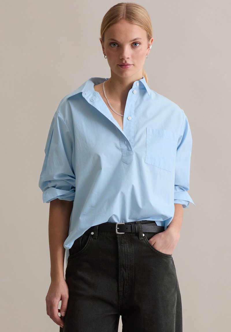 Hellblaues, übergroßes Hemd mit Kragen und einer Brusttasche, kombiniert mit dunkelgrünen Jeans und einem schwarzen Gürtel, mit hochgekrempelten Ärmeln.