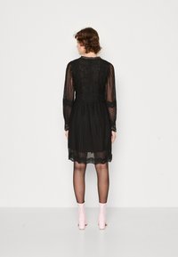 Schwarzes Spitzenkleid mit langen Ärmeln, strukturiertem Design, tailliertem Schnitt und einem Saum mit Spitzenrand, kombiniert mit zarten rosa Stiefeletten.