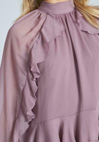 Blusa viola chiaro con colletto alto, dettagli arricciati lungo le spalle e tessuto testurizzato che crea una silhouette morbida e fluida.