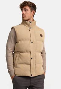 Beige Corduroy-Puffer-Weste mit hochgeschlossenem Kragen, vorderen Knöpfen, zwei Seitentaschen und strukturierter Stoffoberfläche. Rundes Logo auf der Brust.