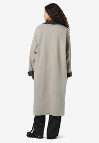 Vrouw met lang krullend haar, gekleed in een beige lange jas met een zwarte kraag en mouwen, zwarte broek en zwarte schoenen, staande met haar rug naar de camera gekeerd.