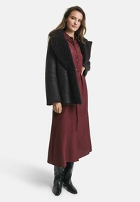 Schwarze Schaffelljacke mit einem breiten Kragen über einem weinroten Langarmkleid. Das Kleid hat eine durchgehende Knopfleiste und eine gebundene Taille. Schwarze kniehohe Stiefel.