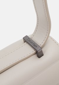 Torebka Calvin Klein w jasnobeżowej skórze, charakteryzująca się gładką teksturą i kontrastowym metalowym akcentem z wygrawerowanym logo na pasku.