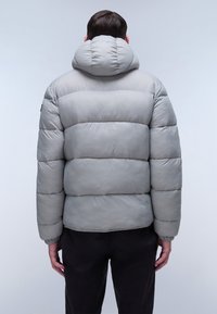 Uomo che indossa un giubbotto imbottito grigio chiaro con cappuccio, visto da dietro, su uno sfondo chiaro e uniforme.