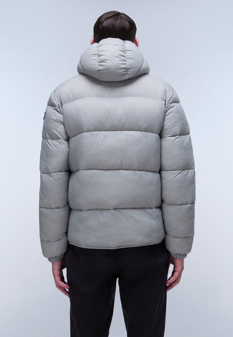 Mann, der eine hellgraue Steppjacke mit Kapuze trägt, von hinten betrachtet, vor einem einfarbigen hellen Hintergrund.