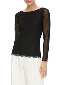 Czarny top z długim rękawem, prześwitującymi rękawami, miękkiego materiału, okrągłym dekoltem i falbaniastym wykończeniem. Połączony z białymi spodniami o szerokich nogawkach.