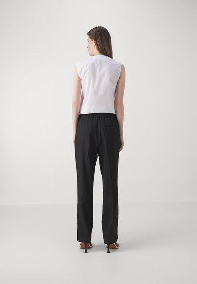 White sleeveless top with a fitted design, paired with black loose pants featuring a back pocket and slits at the hem, completed with heeled shoes.