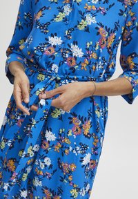 Rochie florală albastră, confecționată dintr-un material ușor. Prezintă un decolteu cu nasturi, mâneci lungi și un talie legată, punând în evidență flori vibrante în diverse culori.