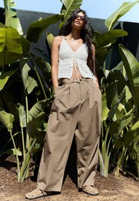 Virkat crop top i vitt med knytning framtill, kombinerad med beigefärgade vida byxor. Sandaler med öppen design. Gröna löv i bakgrunden.